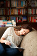 © Marine Gastineau/peopleimages.com - Calm, library and woman reading a book on a sofa for literature while relaxing with shelves. Rest, happy and female person with a smile enjoying a fantasy story or novel on couch in bookstore or shop
