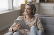 © fizkes - Happy dreamy mature woman holding tablet, sitting on comfortable home couch, looking at window away, smiling, laughing, thinking, dreaming, planning, using wireless Internet technology