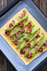 Poster - Uncooked puff pastry, green asparagus with mozzarella cheese, mushrooms and cherry tomatoes on baking paper on cooking pan
