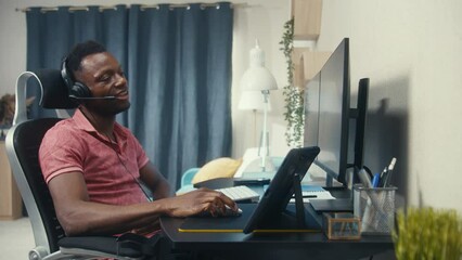 African American web designer or developer works remotely at home. Man sits and talks on online conference at computer. Tablet and multiple monitors stands on table. Concept of working at cozy home.