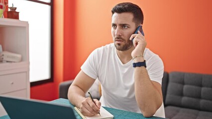 Wall Mural - Young hispanic man talking on smartphone using laptop taking notes at dinning room