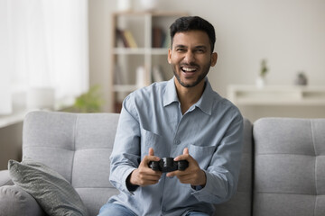 Joyful young Indian gamer guy playing videogame at home, holding gamepad, joystick, game controller, laughing, shouting, sitting on couch, looking at camera, enjoying leisure, virtual activity