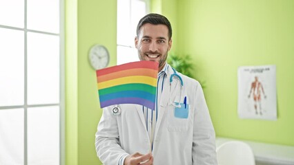 Wall Mural - Young hispanic man doctor smiling confident holding lgbtq flag at clinic