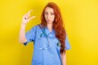 young red-haired doctor woman over yellow studio background purses lip and gestures with hand, shows something very little.