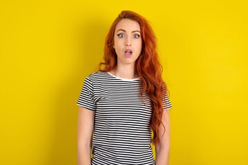 Wall Mural - Shocked young beautiful red haired woman wearing striped t shirt over yellow studio background stares bugged eyes keeps mouth opened has surprised expression. Omg concept