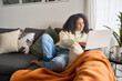 © insta_photos - Latin young woman sitting on couch using laptop at home surfing, doing ecommerce shopping, looking at computer communicating online, searching on web, watching videos or elearning.