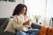 Leinwandbild Motiv Happy young latin woman sitting on sofa holding mobile phone using cellphone technology doing ecommerce shopping, buying online, texting messages relaxing on couch in cozy living room at home.