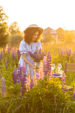 Golden Hour, Sunset, Summer. Beautiful Happy Young Woman On Meadow Arranging Table For Outdoor Event, Gathering Wildflowers. Wedding Or Romantic Date Decoration In The Field With Purple Lupins