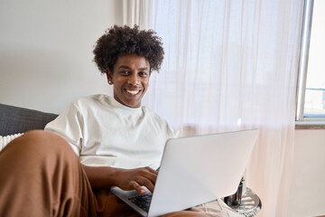 Wall Mural - Happy smiling African American gen z teen student sitting at couch relaxing at home with laptop computer looking at camera advertising online learning, remote web elearning. Authentic portrait.