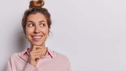 Wall Mural - Studio shot of beautiful woman smiles toothily has gentle expression smiles tenderly keeps hand under chin concentrated wears shirt isolated on white background copy space for your advertising content