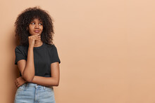 Horizontal Shot Of Curly Haired Woman Captured In Moment Of Deep Contemplation Gazes Into Distance Evoking Sense Of Curiosity And Wonder Keeps Hand On Chin Wears Black T Shirt Jeans Isolated On Beige
