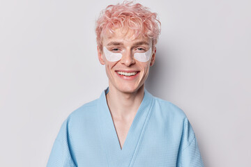 Wall Mural - Cheerful adult European man with pink hairstyle applies cosmetic patches his toothy smile reflects enthusiasm dressed in cozy bathrobe indulges in his morning routine isolated on white background.