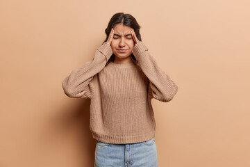 Iranian woman holds her temples her eyes closed tightly in attempt to alleviate pounding headache dressed casually and isolated against beige background expresses sense of desperation and frustration