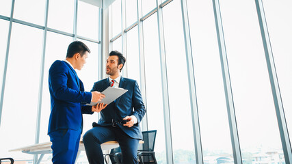 Two business people talk project strategy at office meeting room. Businessman discuss project planning with colleague at modern workplace while having conversation and advice on financial report. Jivy