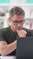 Wall Mural - 50s businessman with gray hair working from home, thinking. Man in casual sitting at desk using laptop computer, business manager online in home office.