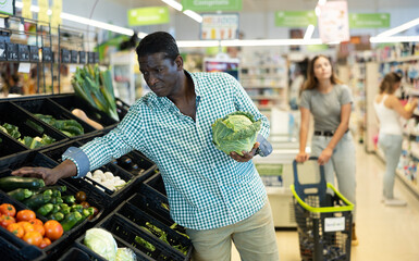 Focused African man choosing cabbage and zucchini near the counter in greengrocery store