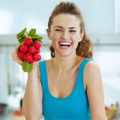 Wall Mural - Smiling young woman holding bunch of radishes in kitchen