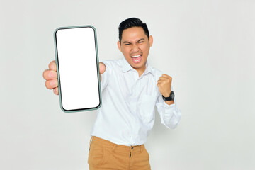 Mockup. Excited young Asian man in formal wear showing mobile phone with blank white screen and celebrating success while clenching fist isolated on white background