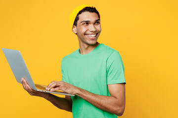 Wall Mural - Side view young IT man of African American ethnicity wears casual clothes green t-shirt hat hold use work on laptop pc computer look aside isolated on plain yellow background studio Lifestyle concept.