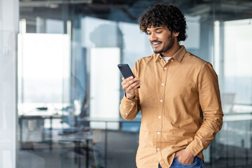 A young Hispanic stands inside office, the man holds the phone in his hands, the businessman browses online pages and uses applications, dials a message, makes a call, a smiling satisfied employee.