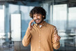 © Liubomir - Young successful hispanic businessman in a shirt talking on the phone inside the office, man standing near the window cheerfully communicating with colleagues and clients.
