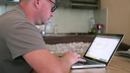 Caucasian man with glasses writes program code on laptop, works at home in computer looking on monitor screen. IT specialist, programmer working at home.