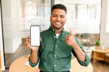 Portrait of handsome young Indian man demonstrates smartphone with blank screen and shows thumb up, guy likes mobile app, freelancer looking and smiling at the camera, feels satisfied
