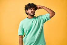 Portrait Of A Handsome Young Man In A Turquoise T-shirt On A Yellow Background, Scratching His Head And Looking Very Pensively To The Side. The Emotion Of Doubt, Reflection And Confusion.