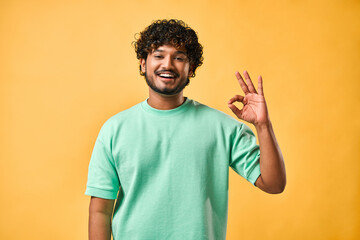 Handsome indian man in turquoise t-shirt showing ok gesture with one hand while looking at camera and smiling while standing against yellow background.