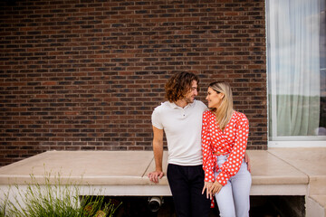 Smiling young couple in love sitting in front of house brick wall