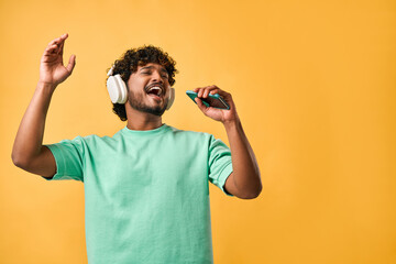 Portrait of handsome curly Indian man in turquoise t-shirt and singing with phone in wireless headphones enjoying music and gesturing emotionally.