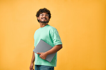 Portrait of handsome curly Indian man in turquoise t-shirt and glasses laughing looking away and holding laptop. Copy space.