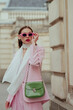 Fashionable elegant confident woman wearing trendy pink sunglasses, suit blazer, white silk scarf, trousers, with green faux leather shoulder bag, posing in street. Outdoor fashion portrait