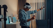© tirachard - Young Asian man gamer wear gray hoodie with VR oculus hold VR joystick controllers feeling exciting with new experience of game stand front sofa living room home at night. Virtual reality concept.