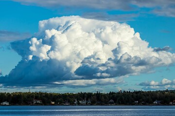 Huge cumulus cloud hanging over a silent blue lake bordered by a line of thick trees on a sunny day