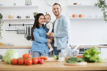 Wall Mural - Portrait of joyful young parents in denim clothes cuddling little daughter while staying in kitchen of spacious apartment. Loving adult people and kid lifting mood while cooking together at home.