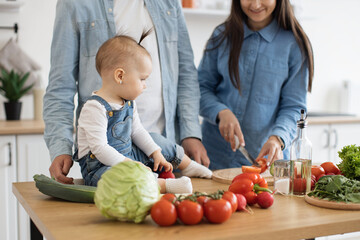 Wall Mural - Cropped view of brunette female holding cucumber while sweet baby girl reaching out hand for green vegetable. Loving young mother teaching daughter eating healthy meal prepared together at home.