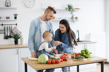 Wall Mural - Caring young woman in denim clothes letting baby girl taste green vegetable while cooking dishes for dinner at noon. Mindful parents getting child used to new textures and new flavours of solid food.