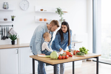 Wall Mural - Funny little girl in cozy outfit sitting on dining table while caring young mother holding bite-sized slice of tomato. Mindful parents motivating infant daughter to making healthier food choices.