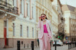 Fashionable elegant confident woman wearing trendy pink sunglasses, suit blazer, satin top, white silk scarf, trousers, walking in street of European city. Copy, empty space for text