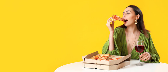 Wall Mural - Sexy young woman with glass of wine eating tasty pizza on yellow background with space for text