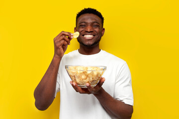 happy african american man holding plate of chips and eating snacks from potatoes on yellow background