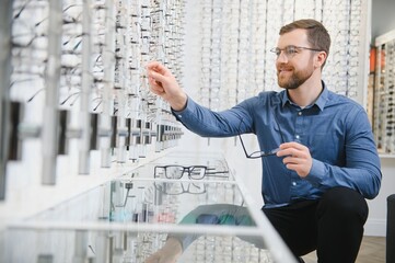 Wall Mural - In Optics Shop. Portrait of male client holding and wearing different spectacles, choosing and trying on new glasses at optical store. Man picking frame for vision correction, closeup.