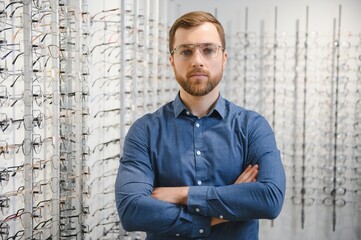 Wall Mural - Satisfied Customer. View of happy young male client wearing new glasses, standing near rack and showcase with eyewear. Smiling man trying on spectacles