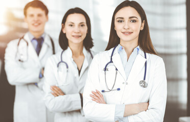 Wall Mural - Group of young professional doctors standing as a team with arms crossed in sunny hospital. Medical help, insurance in health care, best disease treatment and medicine concept