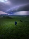Fototapeta  - Vertical shot of a hiker on a grassy pasture looking at lightning in the distance