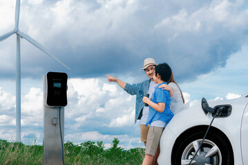 Concept of progressive happy family enjoying their time at wind farm with electric vehicle. Electric vehicle driven by clean renewable energy from wind turbine generator for charging station.