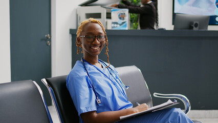 Wall Mural - Female nurse looking at medical reports in waiting area, sitting in facility lobby. Young healthcare assistant looking at checkup forms before having consultation appointment in health center.