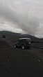 Vertical video. Yellow retro colorful jeep SUV riding black volcano lava field dirt dust desert at mountain aerial view. FPV drone shot speed automobile rally safari travel extreme sport auto drive 4k