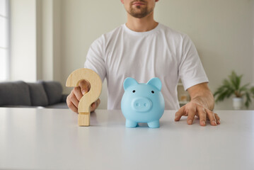 Cropped shot of man sitting at white desk with blue piggy bank and wooden question mark interrogation sign. Closeup objects, close up view. Money, financial uncertainty, confusion, crisis concept